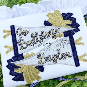 以下が含まれることがあります： 白いTシャツにペナントのデザイン。ペナントには「Go Bulldogs」と「Baylor」の文字が金色の筆記体で書かれており、ネイビーブルーとゴールドのアクセントが付いています。デザインには金色のリボンとネイビーブルーのアクセントが含まれています。