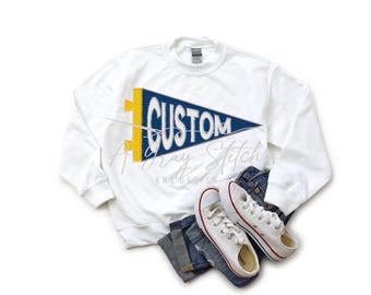 May include: White sweatshirt with a blue and yellow pennant design that says "CUSTOM". The sweatshirt is laying on a pair of blue jeans and white sneakers.