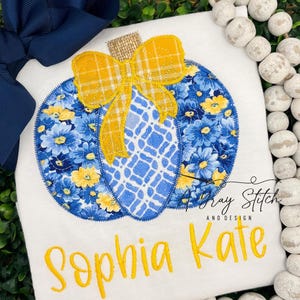 May include: A white t-shirt with a blue and yellow pumpkin applique. The pumpkin has a yellow plaid bow and the text "Sophia Kate" is embroidered in yellow below the pumpkin.