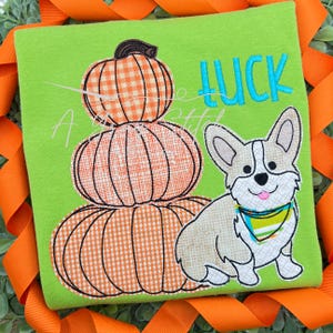 Puede incluir: Una camiseta verde con un diseño bordado de una pila de tres calabazas naranjas y un perro corgi blanco y marrón que lleva un pañuelo azul y verde. El texto "LUCK" está bordado en azul sobre el perro.
