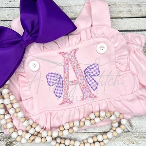 May include: Pink baby romper with a large embroidered letter A decorated with floral and gingham bows. The romper has a ruffled neckline and straps.