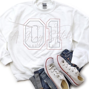 Może przedstawiać: Biała bluza z nadrukowaną czerwoną i niebieską ramką wokół cyfry "01" z przodu. Bluza leży na niebieskich dżinsach i białych butach sportowych.