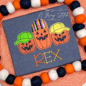 May include: A gray t-shirt with three embroidered pumpkin faces wearing hats. The pumpkins are orange with black eyes, noses, and mouths. The hats are green, yellow, and striped. The name "REX" is embroidered below the pumpkins.