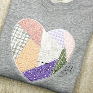 Może przedstawiać: Szara bluza z dużym, pikowanym wzorem serca w odcieniach różowego, fioletowego, zielonego i beżowego. Serce składa się z różnych kawałków tkaniny zszytych ze sobą. Tekst "A Gray Stitch and Design" jest nadrukowany na biało w prawym dolnym rogu bluzy.