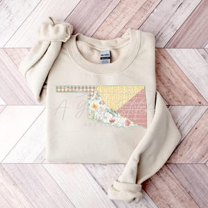 May include: A beige sweatshirt with a patchwork design of the state of Oklahoma. The design features floral patterns, yellow and pink fabric, and the text "A girl and her Oklahoma".