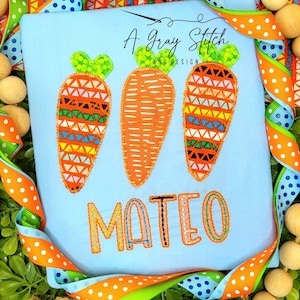 May include: A light blue t-shirt with three embroidered carrots and the name "MATEO" in orange and black thread. The carrots have a geometric pattern and green tops.