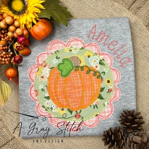 May include: A gray t-shirt with a pink and white scalloped circle design featuring a stitched pumpkin with a green stem and leaves. The text "Amelia" is embroidered in white on the shirt. The text "A Gray Stitch and Design" is embroidered in gray at the bottom of the shirt.