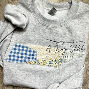 Könnte beinhalten: Ein graues Sweatshirt mit einem Patchwork-Design aus blauem und gelbem Gingham und Blumenstoff in Form des Bundesstaates Tennessee. Der Text "A Gray Stitch and Design" ist auf das Sweatshirt gestickt.