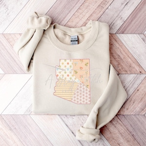May include: A beige sweatshirt with a patchwork design of the state of Arizona. The design is in shades of pink, yellow, and white.