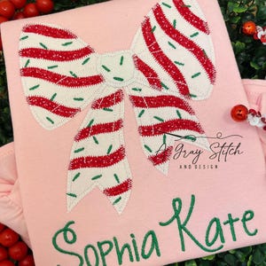 May include: Pink shirt with a red and white striped bow applique. The bow has green sprinkles. The shirt is personalized with the names "Sophia Kate" in green embroidery.