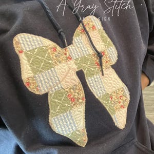 Peut inclure: Un sweat-shirt noir avec un grand nœud en patchwork cousu sur le devant. Le nœud est fait de différents tissus dans des tons de vert, bleu et blanc. Le texte "A Gray Stitch" est imprimé sur le sweat-shirt.