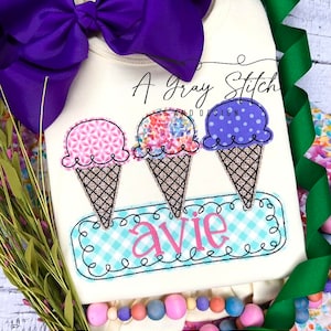 May include: A white onesie with a design of three ice cream cones with different colored scoops and a blue and white checkered banner with the name "Avie" embroidered on it.