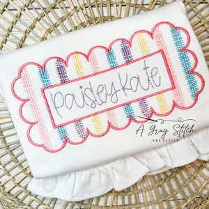May include: White fabric with a pink, yellow, blue, and purple striped design. The design is framed with a pink scalloped border. The name "paisley kate" is embroidered in purple inside a pink rectangle.