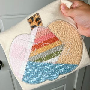 Puede incluir: Una camiseta beige con un diseño de calabaza de patchwork colorido. La calabaza está hecha de diferentes telas con varias texturas y patrones, incluyendo estampado de leopardo, rayas y lunares.