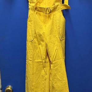 Peut inclure: Une combinaison jaune vif avec des manches courtes, un col chemise et une taille ceinturée. La combinaison comporte une fermeture éclair, des poches avant et un pantalon large. Une étiquette jaune est suspendue à la ceinture.