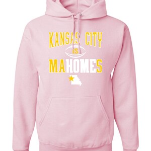Puede incluir: Sudadera con capucha rosa con un gr&aacute;fico blanco y dorado que dice "Kansas City is Mahomes" con un bal&oacute;n de f&uacute;tbol y una estrella sobre el estado de Misuri.