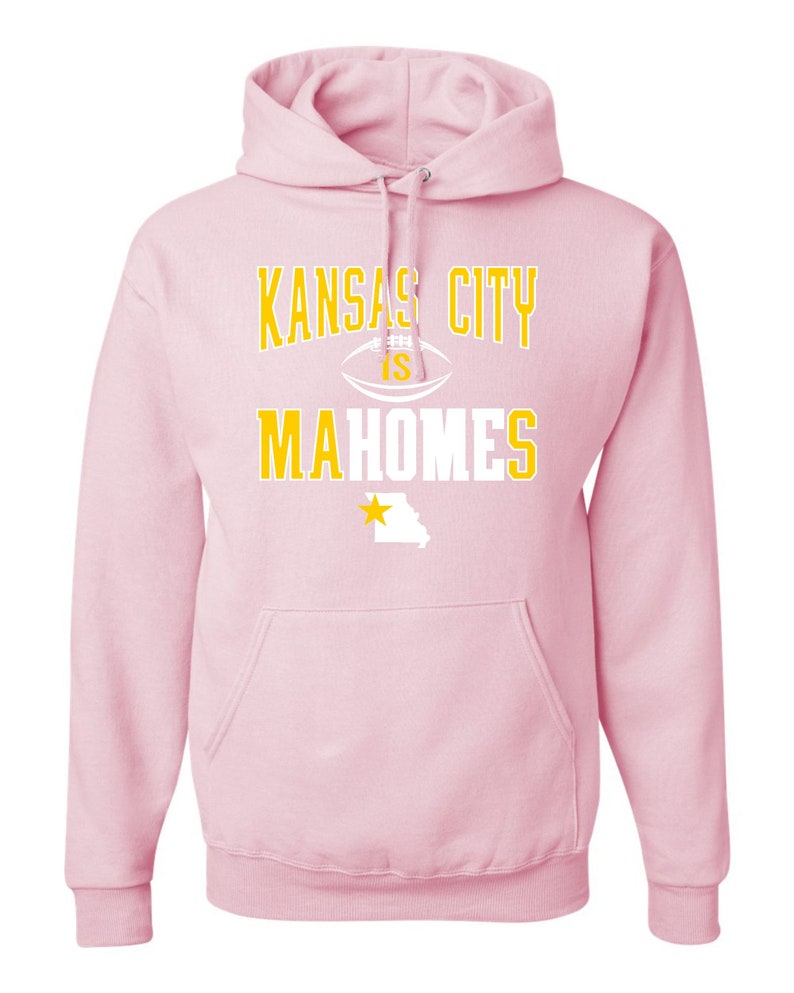 Puede incluir: Sudadera con capucha rosa con un gr&aacute;fico blanco y dorado que dice "Kansas City is Mahomes" con un bal&oacute;n de f&uacute;tbol y una estrella sobre el estado de Misuri.