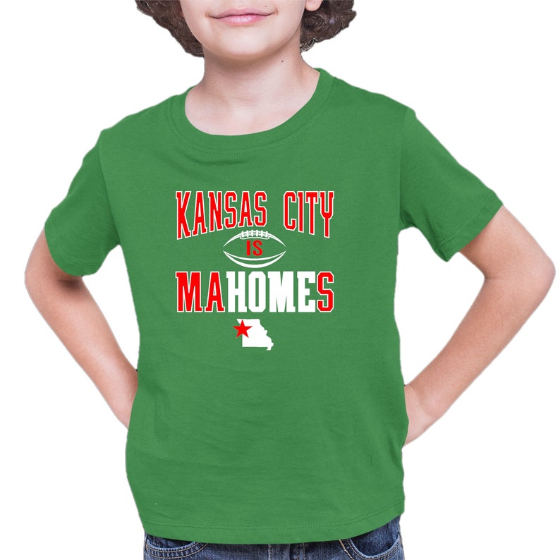 K&ouml;nnte beinhalten: Gr&uuml;nes T-Shirt mit einer wei&szlig;en Grafik, auf der "Kansas City is Mahomes" steht, mit einem Football und einem roten Stern &uuml;ber einem wei&szlig;en Umriss des Bundesstaates Missouri.