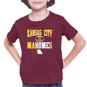 K&ouml;nnte beinhalten: Ein maroonfarbenes T-Shirt mit einer wei&szlig;en Grafik, auf der "Kansas City is Mahomes" steht, mit einem Fu&szlig;ball und einem gelben Stern &uuml;ber dem Bundesstaat Missouri.