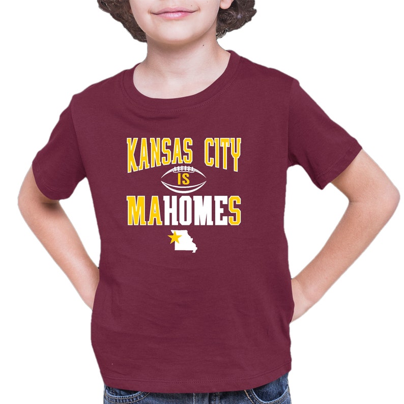 K&ouml;nnte beinhalten: Ein maroonfarbenes T-Shirt mit einer wei&szlig;en Grafik, auf der "Kansas City is Mahomes" steht, mit einem Fu&szlig;ball und einem gelben Stern &uuml;ber dem Bundesstaat Missouri.