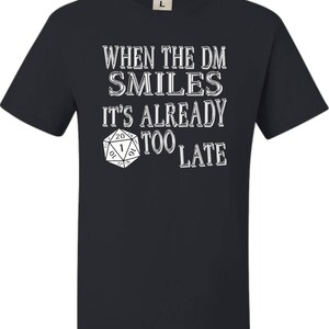 May include: Black t-shirt with white text that reads "When the DM smiles it's already too late". A white 20-sided die is pictured below the text.