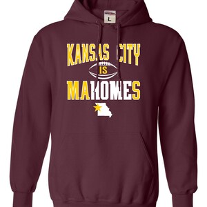 Puede incluir: Sudadera con capucha color granate con el texto "Kansas City is Mahomes" en letras blancas y amarillas. Una estrella amarilla y el contorno del estado de Misuri se encuentran debajo del texto.