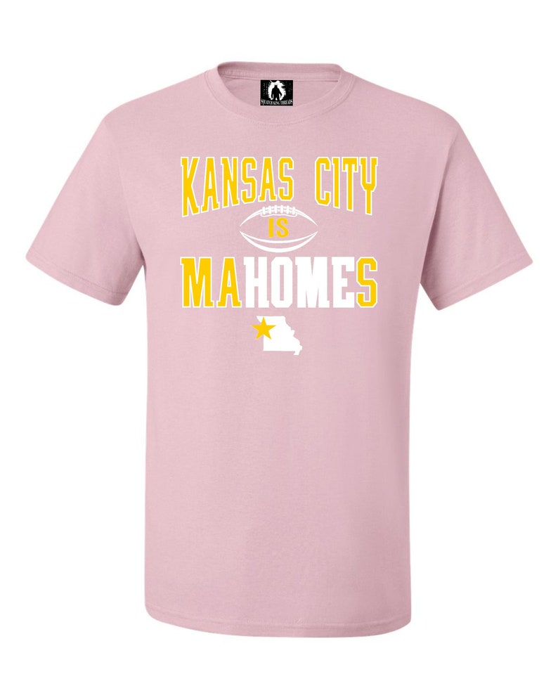 K&ouml;nnte beinhalten: Rosafarbenes T-Shirt mit einer wei&szlig;en Grafik, auf der "Kansas City is Mahomes" steht, mit einem Fu&szlig;ball und einem gelben Stern &uuml;ber dem Bundesstaat Missouri.