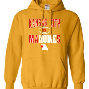 Puede incluir: Una sudadera con capucha amarilla con un gr&aacute;fico blanco que dice "Kansas City is Mahomes" en letras rojas. El gr&aacute;fico tambi&eacute;n incluye un bal&oacute;n de f&uacute;tbol y una estrella roja sobre el contorno del estado de Misuri.