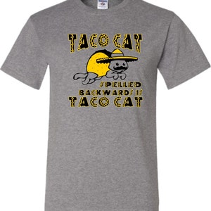 以下が含まれることがあります： グレーのTシャツに、黒い帽子とタコスシェルを被った猫の黒と黄色のイラストが描かれています。猫の上に「Taco Cat」という文字、猫の下に「Spelled backwards is Taco Cat」という文字が書かれています。