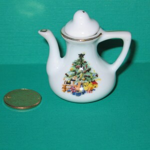 May include: White porcelain teapot with a Christmas tree design. The teapot has a gold trim and a handle. The design features a Christmas tree with presents and a star on top.