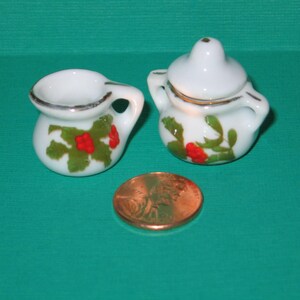 May include: Two miniature white ceramic pitchers with green and red floral designs. The pitcher on the left has a handle and a wide opening. The pitcher on the right has a lid and a smaller opening.