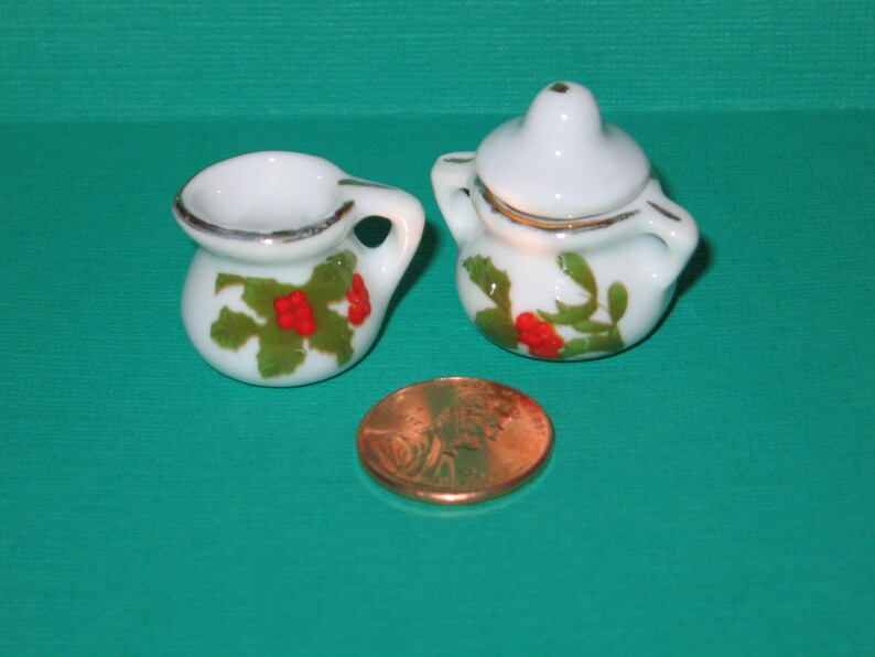 May include: Two miniature white ceramic pitchers with green and red floral designs. The pitcher on the left has a handle and a wide opening. The pitcher on the right has a lid and a smaller opening.