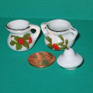 May include: Two white ceramic teacups with green and red holly designs. The cups have a silver rim and are on a green surface.