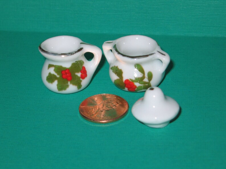 May include: Two white ceramic teacups with green and red holly designs. The cups have a silver rim and are on a green surface.