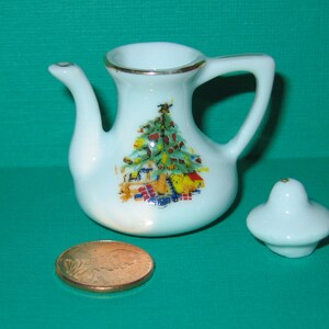 May include: A white porcelain teapot with a gold trim and a Christmas tree design. The teapot has a lid and a handle. The teapot is sitting on a green surface.