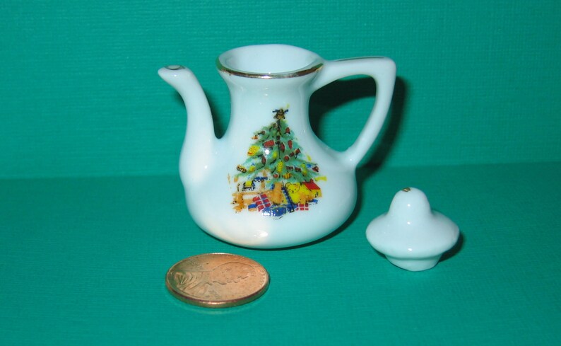 May include: A white porcelain teapot with a gold trim and a Christmas tree design. The teapot has a lid and a handle. The teapot is sitting on a green surface.