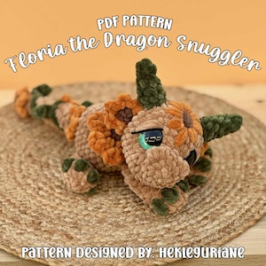 Op de afbeelding: Een gehaakt drakenfiguur genaamd Floria the Dragon Snuggler, met beige, donkergroene en oranje garentexturen, grote zwarte en groene ogen en een lange staart. De tekst PDF PATTERN Floria the Dragon Snuggler en PATTERN DESIGNED BY: HEKLEGURIANE is zichtbaar.