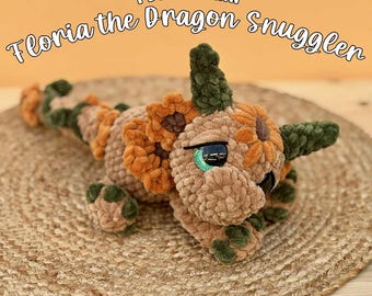 May include: A crochet dragon plushie named Floria the Dragon Snuggler, featuring tan, orange, and dark green yarn. The toy has a textured body, a long tail, and large, dark, glossy eyes. Text reads: PDF PATTERN Floria the Dragon Snuggler PATTERN DESIGNED BY: HEKIEGURIANE.