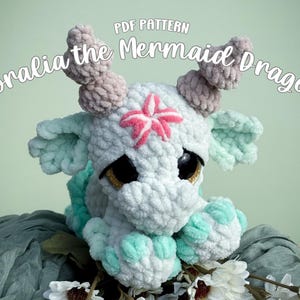 Op de afbeelding: Een handgemaakt gehaakt draakspeeltje, met een witte body met lichtblauwe en roze accenten. De draak heeft grote, expressieve ogen en is versierd met een roze bloem. De tekst "PDF Pattern Coralia the Mermaid Dragon" staat bovenaan.