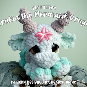 May include: A crochet plush dragon toy with white and mint green yarn, featuring large black eyes, two tan horns, and a pink starfish detail on its forehead. The text reads PDF PATTERN Coralia the Mermaid Dragon and PATTERN DESIGNED BY: HEKLEGURIANE.