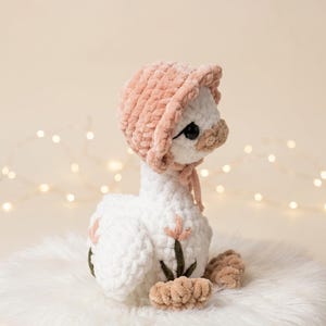 May include: A handmade white plush goose wearing a pink bonnet and tan feet. The goose has black eyes and a tan beak. The plush toy is sitting on a white fluffy surface. The background is a soft beige with blurred lights.