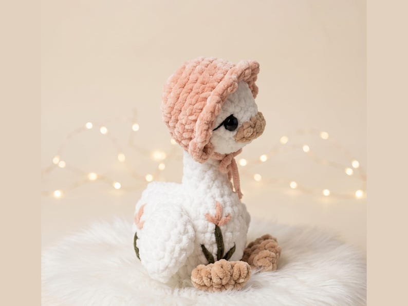 May include: A handmade white plush goose wearing a pink bonnet and tan feet. The goose has black eyes and a tan beak. The plush toy is sitting on a white fluffy surface. The background is a soft beige with blurred lights.