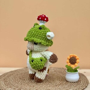 May include: A handmade crochet cow figurine wearing a green mushroom hat and a green bag. The cow has brown spots and a white body. A small crochet sunflower in a white pot is also present.