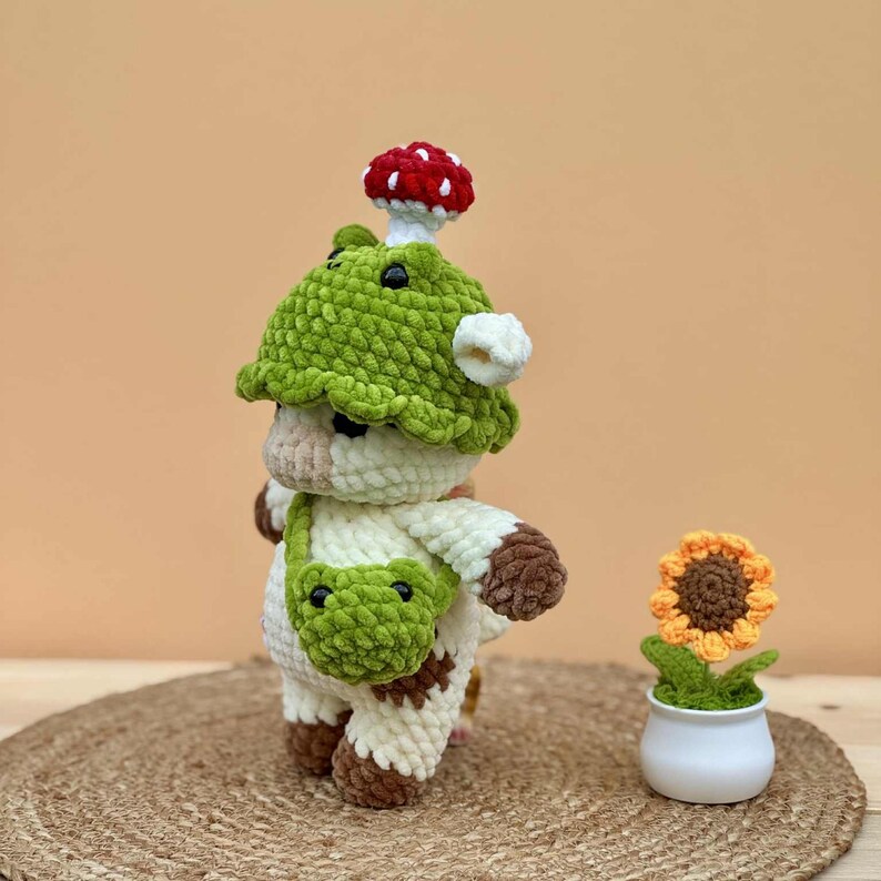 May include: A handmade crochet cow figurine wearing a green mushroom hat and a green bag. The cow has brown spots and a white body. A small crochet sunflower in a white pot is also present.