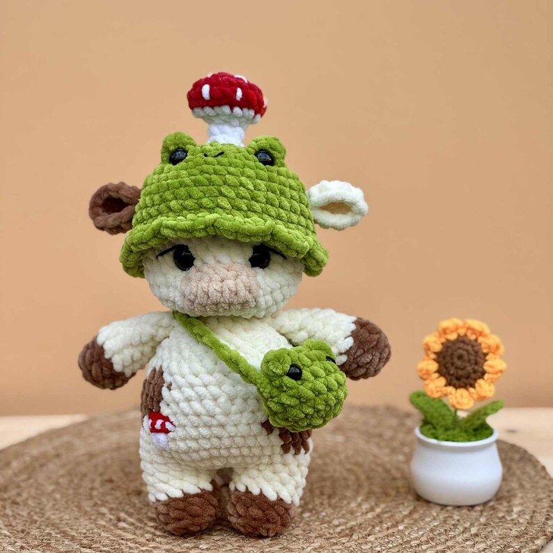 May include: A handmade crochet cow doll with a green frog hat and a mushroom on top. The cow has brown spots and a green frog purse. A small sunflower in a white pot is next to the cow.