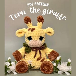 Pode incluir: Um brinquedo de girafa de pelúcia feito à mão em tons de amarelo e marrom, com olhos azuis e um cachecol verde. O texto "Fern the giraffe" e "PDF Pattern" são exibidos na imagem. A girafa é decorada com uma flor branca.