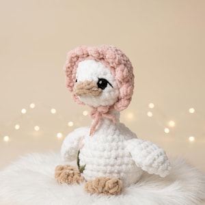 May include: A handmade white crochet goose wearing a pink bonnet with a matching bow. The goose has a tan beak and feet, and black eyes. It is sitting on a fluffy white surface, with soft lighting in the background.