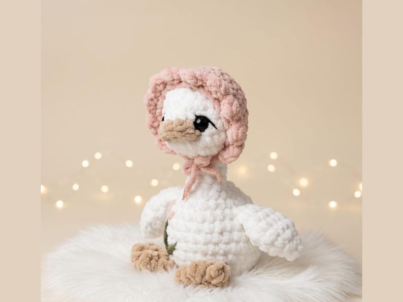 May include: A handmade white crochet goose wearing a pink bonnet with a matching bow. The goose has a tan beak and feet, and black eyes. It is sitting on a fluffy white surface, with soft lighting in the background.