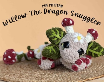 Puede incluir: Un juguete de dragón tejido con características de hongo. El dragón es principalmente beige con detalles de hongos rojos y blancos, alas verdes en forma de hoja y ojos negros grandes y brillantes. El texto "Willow The Dragon Snuggler" está arriba.