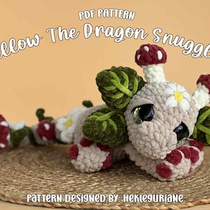 May include: A crochet dragon plushie named Willow The Dragon Snuggler, featuring a beige body, dark green leaves, red and white mushroom accents, and a white daisy on its head. The text PDF PATTERN and PATTERN DESIGNED BY: HEKLEGURIANE are visible.